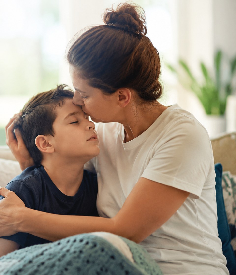 A mother holding her son close and kissing his forehead as he rests against her